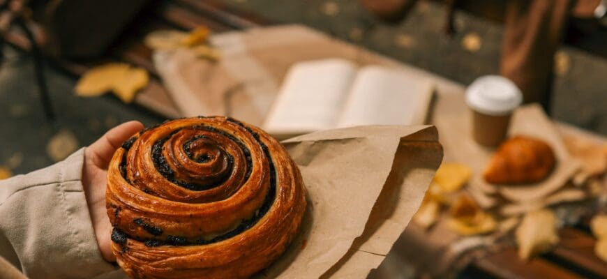 A person holding a morning bun pastry with a mug and a book an a bench in the background